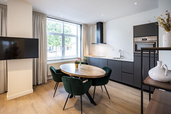 A modern kitchen with a dining area, featuring a round wooden table, green chairs, and a flat-screen TV. - Hotel Botterweck (Valkenburg aan de Geul)