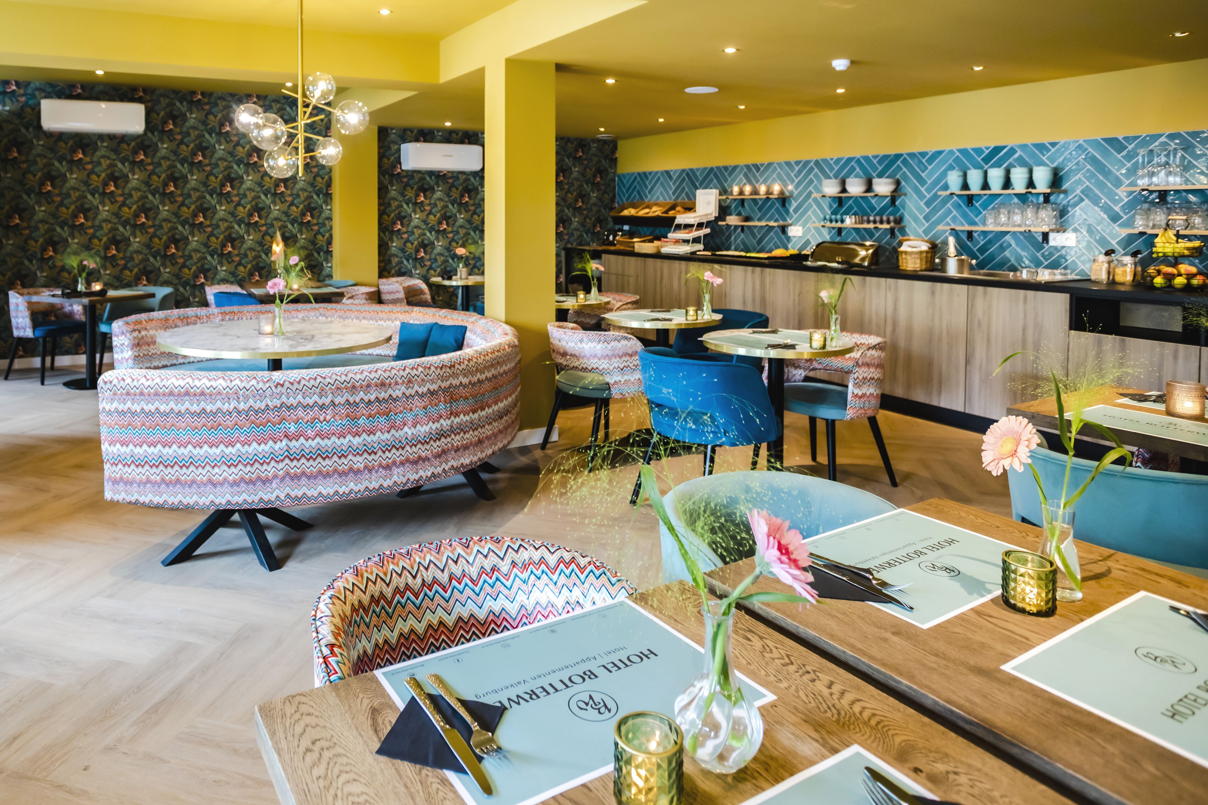 Un restaurante con mesas redondas, asientos estampados y una zona de bar con estanterías llenas de vajilla y copas.
