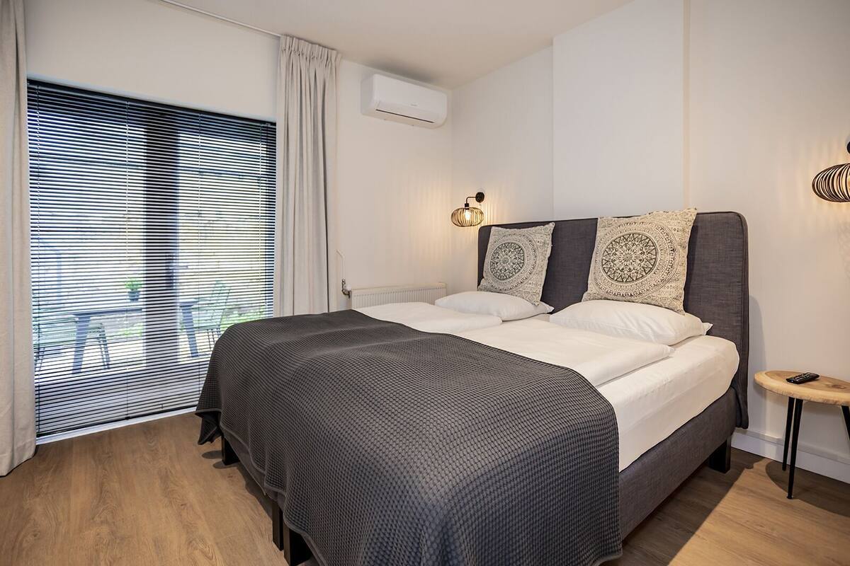 A modern bedroom with a large bed, a bedside table, a wall-mounted air conditioner, and a window with blinds.