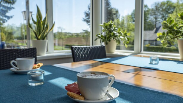 Une table avec une tasse de café, un verre d’eau et un biscuit sur une nappe bleue. On aperçoit, à l’arrière-plan, des plantes en pot et une chaise en osier.