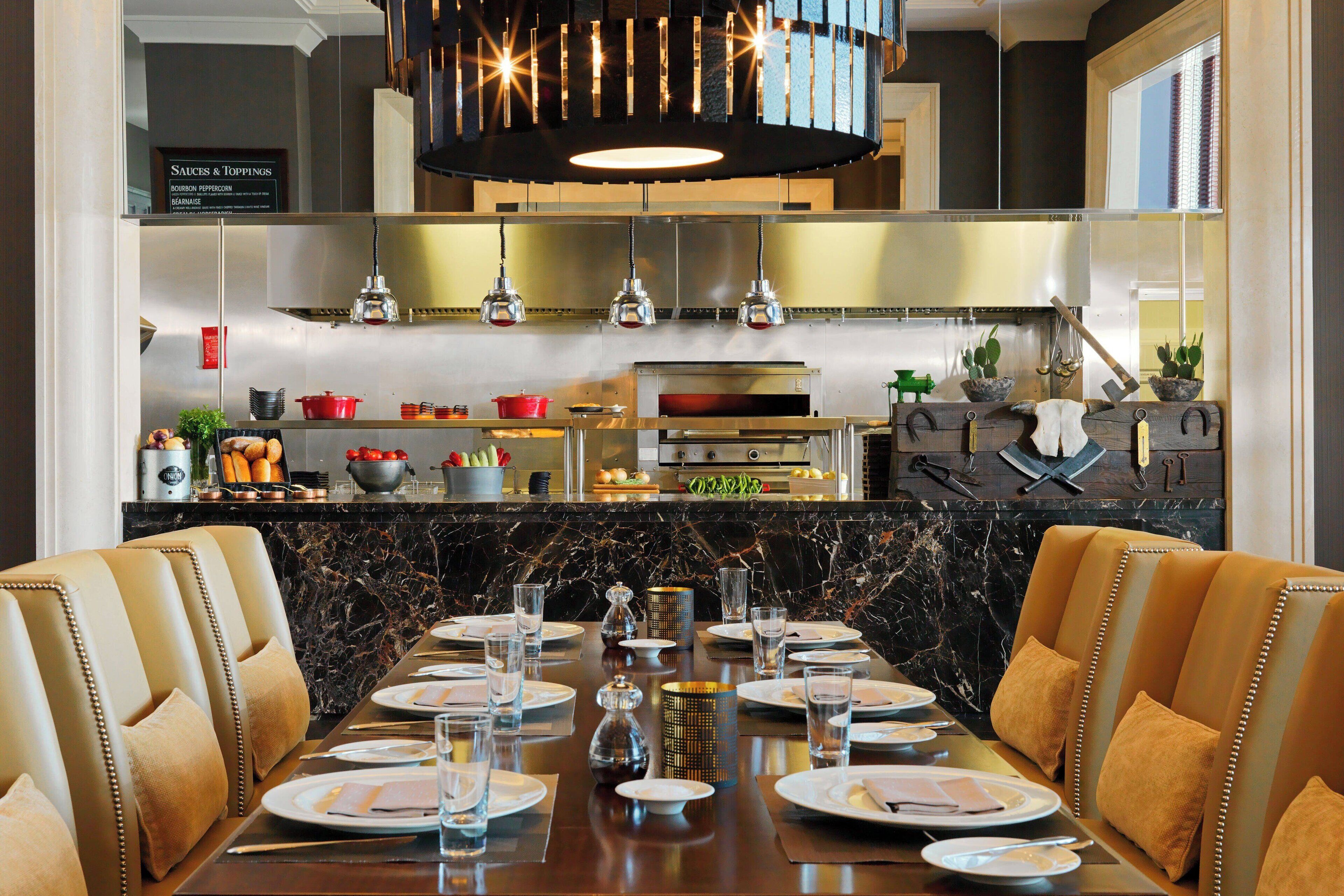 Een restaurant met een lange eettafel gedekt voor meerdere gasten, een keuken op de achtergrond, en een bord waarop sauzen en toppings staan aangegeven.