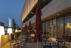 An outdoor dining area with tables and chairs, red umbrellas, and a cityscape view in the background.