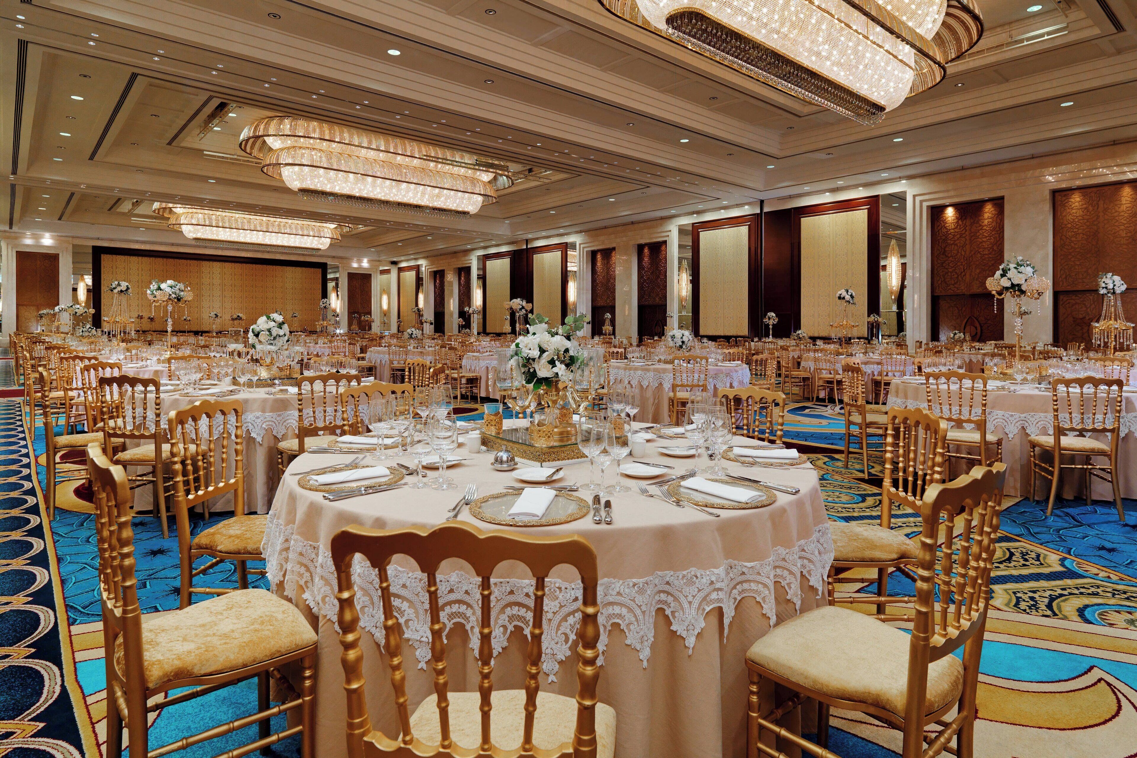 A banquet hall with round tables, gold chairs, and chandeliers.
