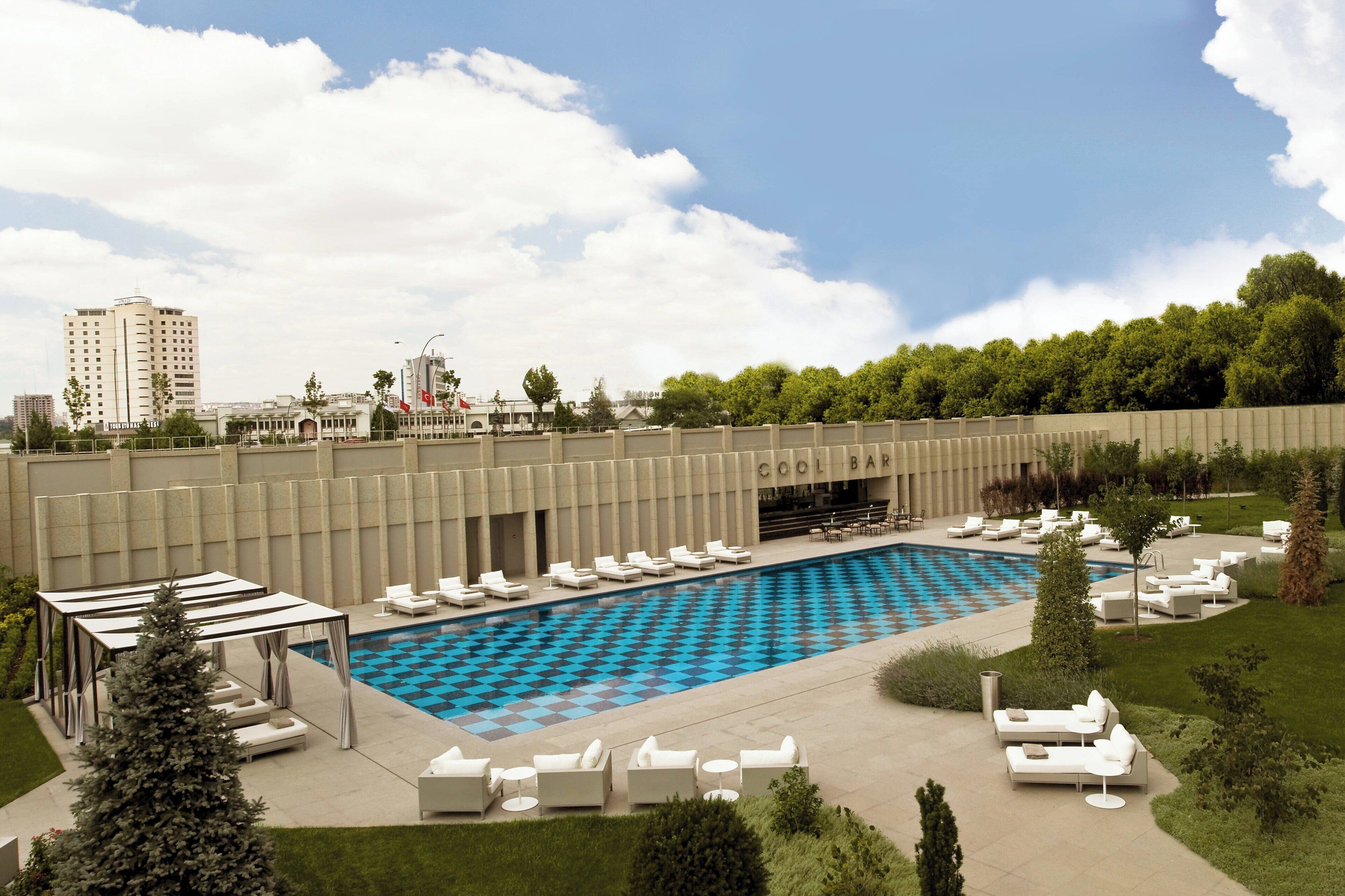 Zona de piscina con sillas de descanso y suelo de ajedrez, rodeada de vegetación y una valla.
