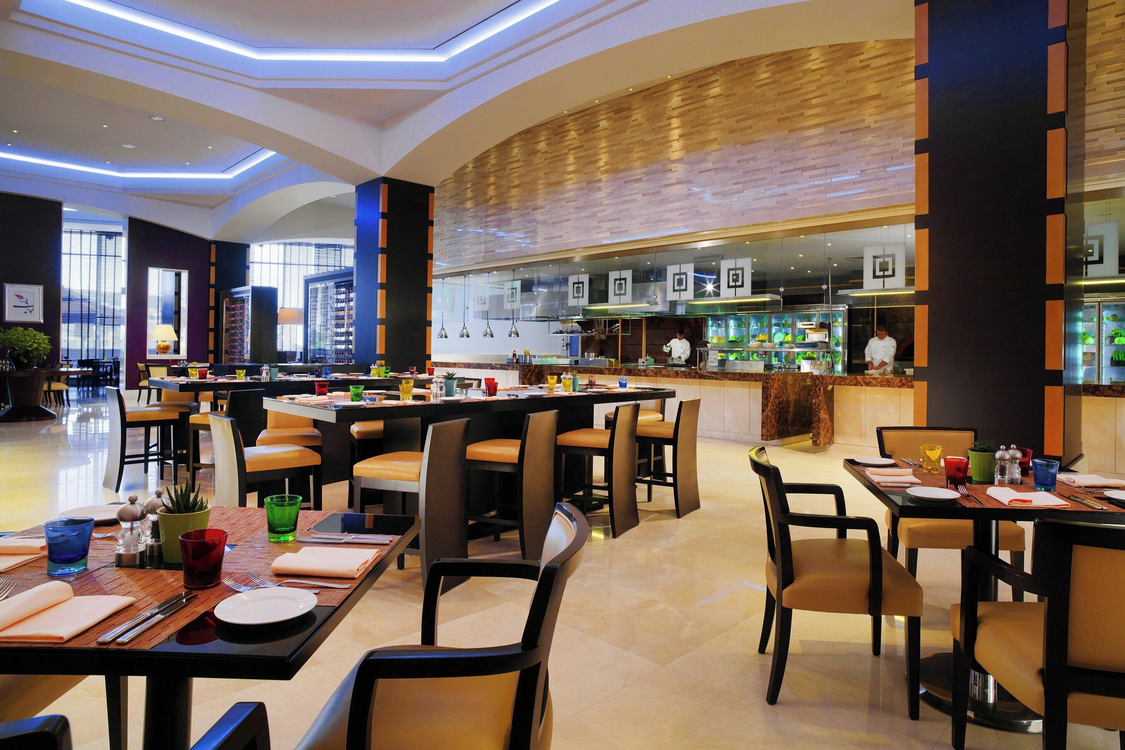 A modern restaurant with multiple dining tables and chairs, a kitchen area, and a view of the cityscape through large windows.