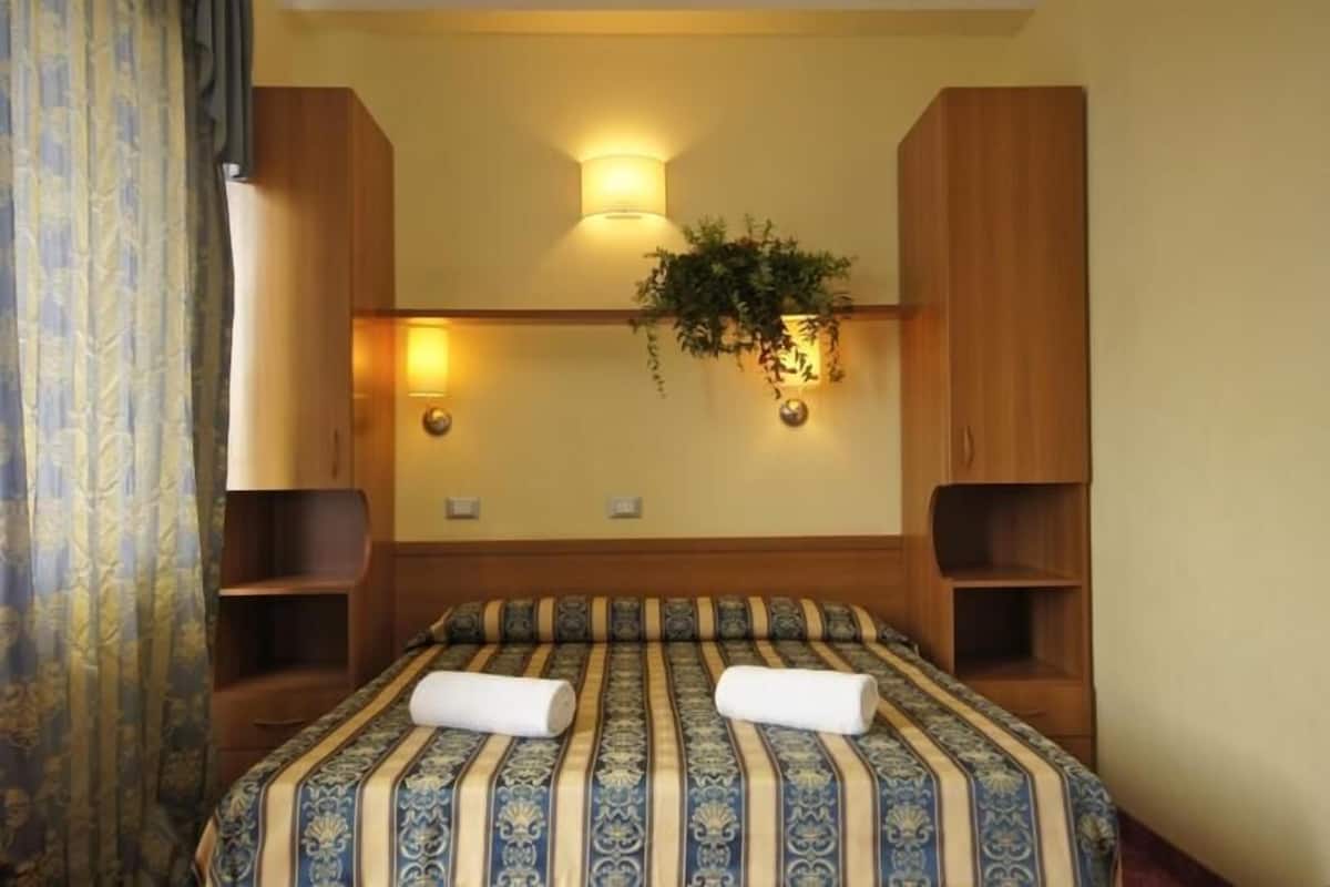 A hotel room with a large bed, wooden headboard, two bedside tables, a wall-mounted plant, and a wall-mounted light fixture.
