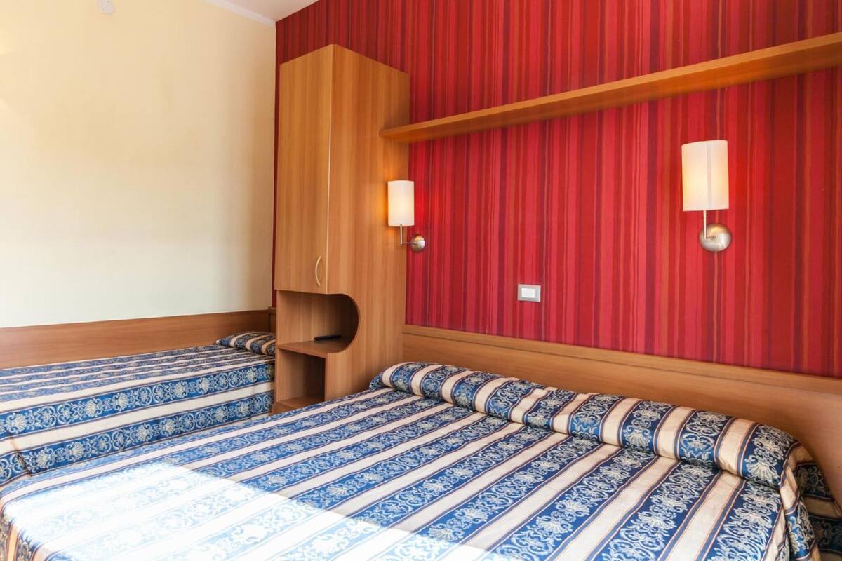 A hotel room with two beds, red striped wallpaper, wooden headboard, and wall-mounted lamps.