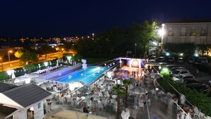 An outdoor pool area with seating and umbrellas, surrounded by trees and a building in the background. - Hotel Promenade Universale (Cesenatico)