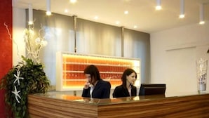 A reception desk with two staff members, a large illuminated display, and a plant to the left. - Hotel Promenade Universale (Cesenatico)