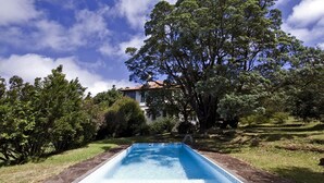 Una piscina rettangolare con bordi in pietra, circondata da vegetazione lussureggiante e alberi, con un edificio sullo sfondo sotto un cielo parzialmente nuvoloso.
