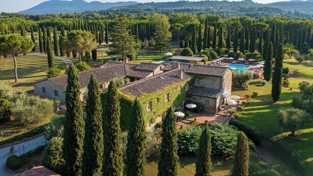 A villa with a swimming pool, surrounded by tall cypress trees and a mountainous backdrop.