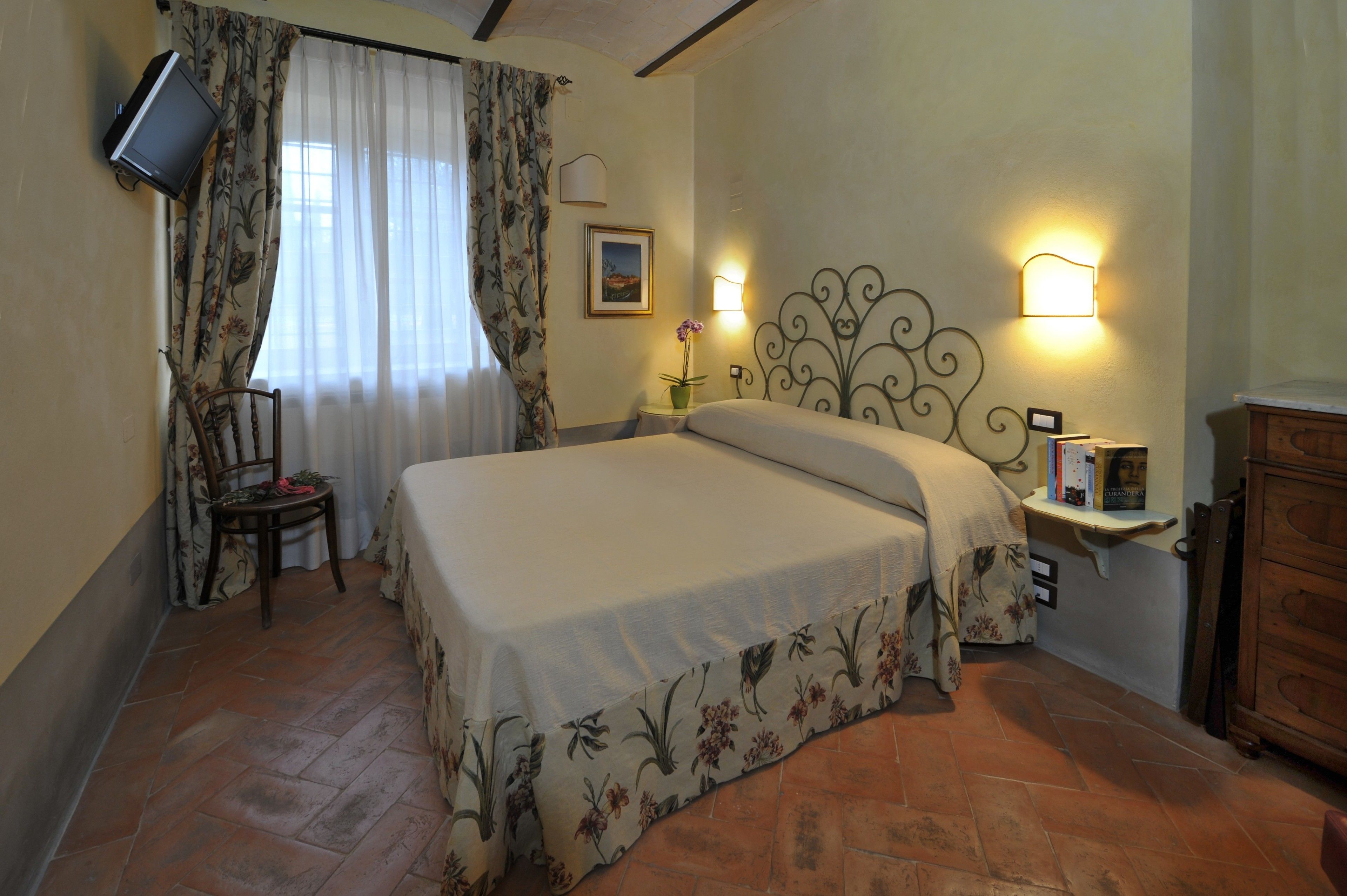 Une chambre à coucher comprenant un lit, une chaise, un téléviseur fixé au mur, une table de chevet avec une lampe et une armoire en bois.