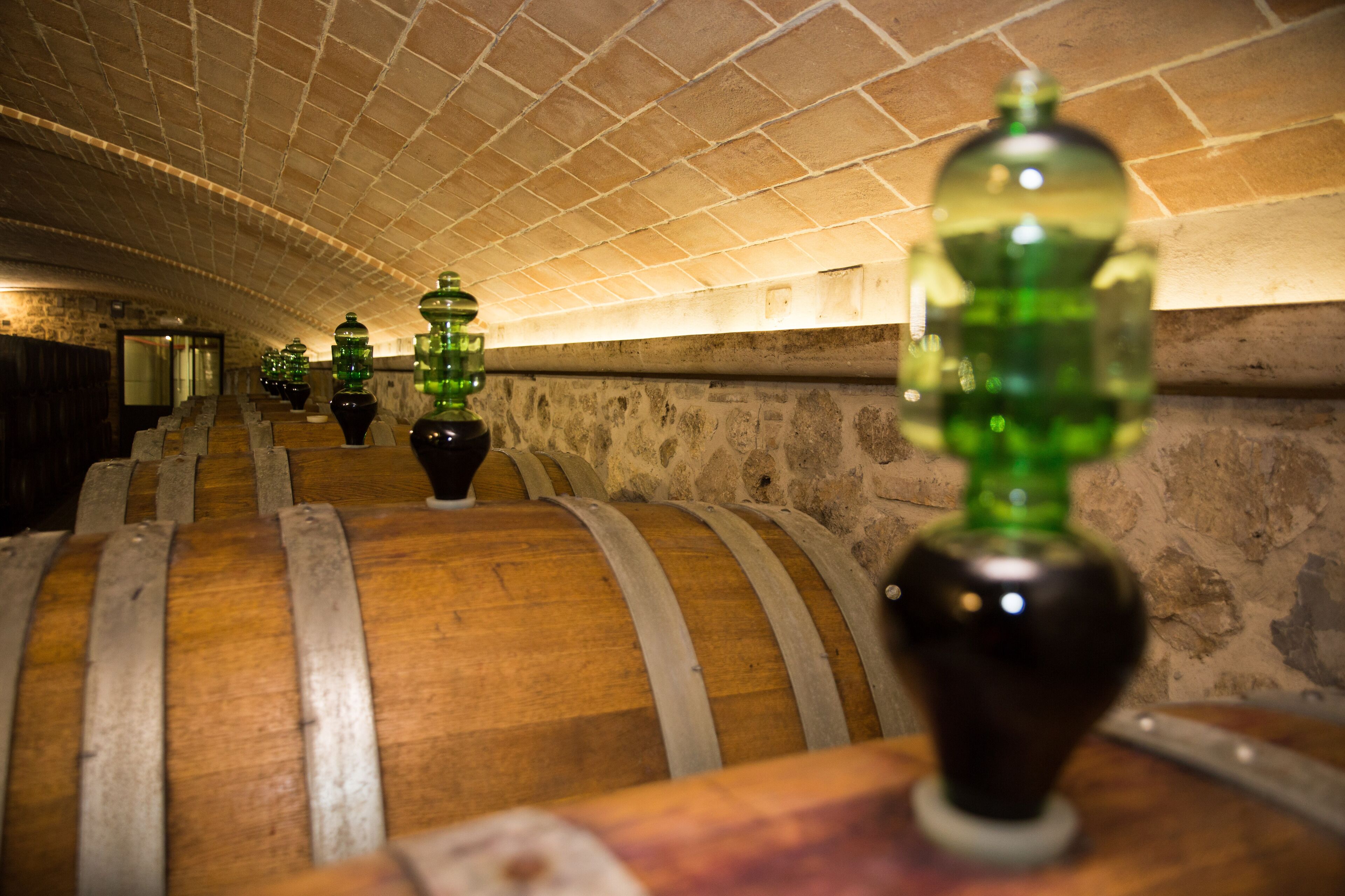 A row of wooden barrels with metal bands, arranged in a dimly lit cellar with stone walls and a brick ceiling.