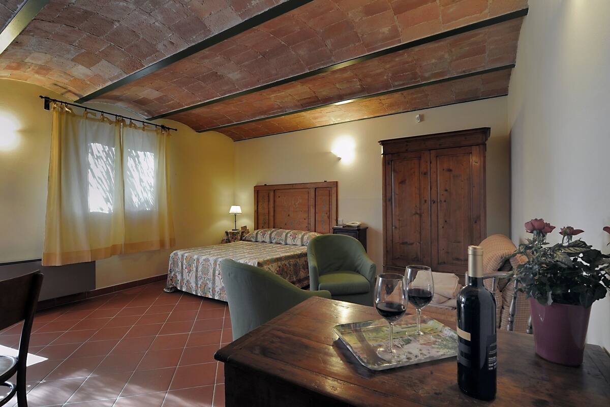 Une pièce confortable avec un lit, une chaise, une table sur laquelle se trouvent une bouteille de vin et des verres, ainsi qu'une plante en pot.