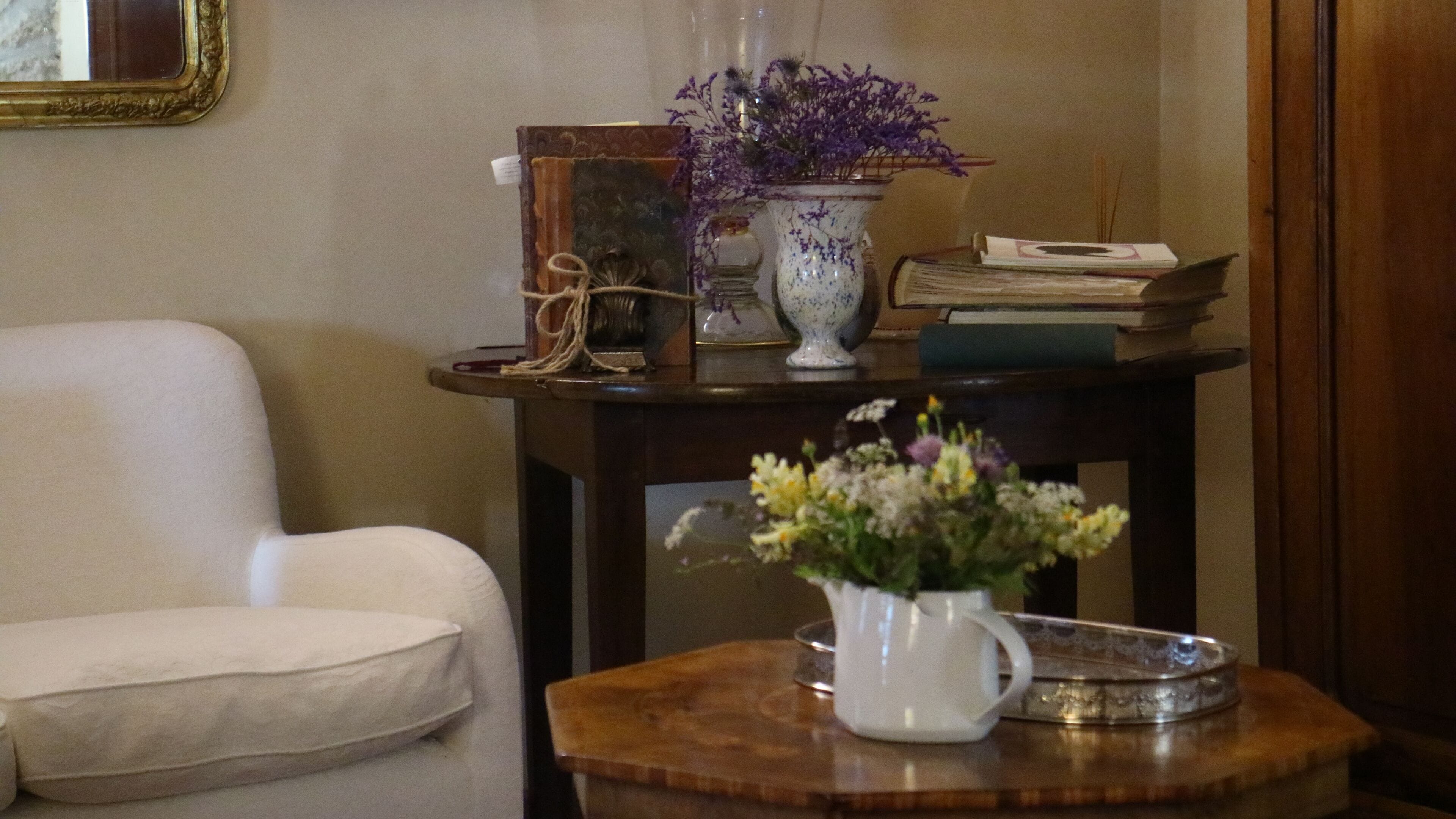 Una habitación con un sillón blanco, una mesita de madera con un jarrón de flores, un libro y un objeto decorativo.