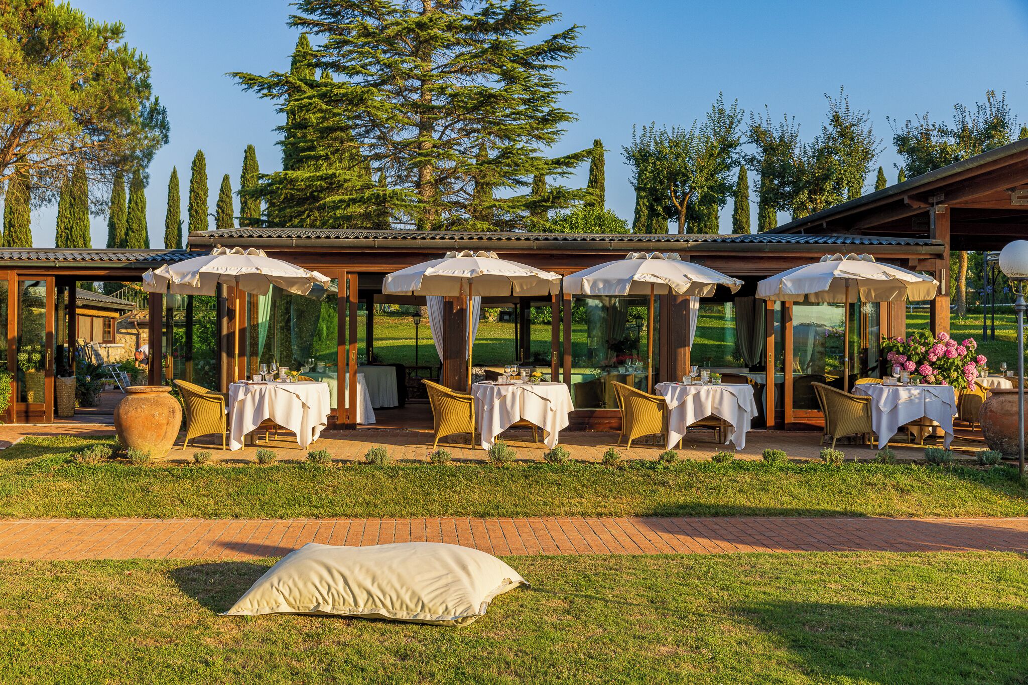 An outdoor restaurant with tables and chairs, covered by umbrellas, set against a backdrop of trees and a clear sky.