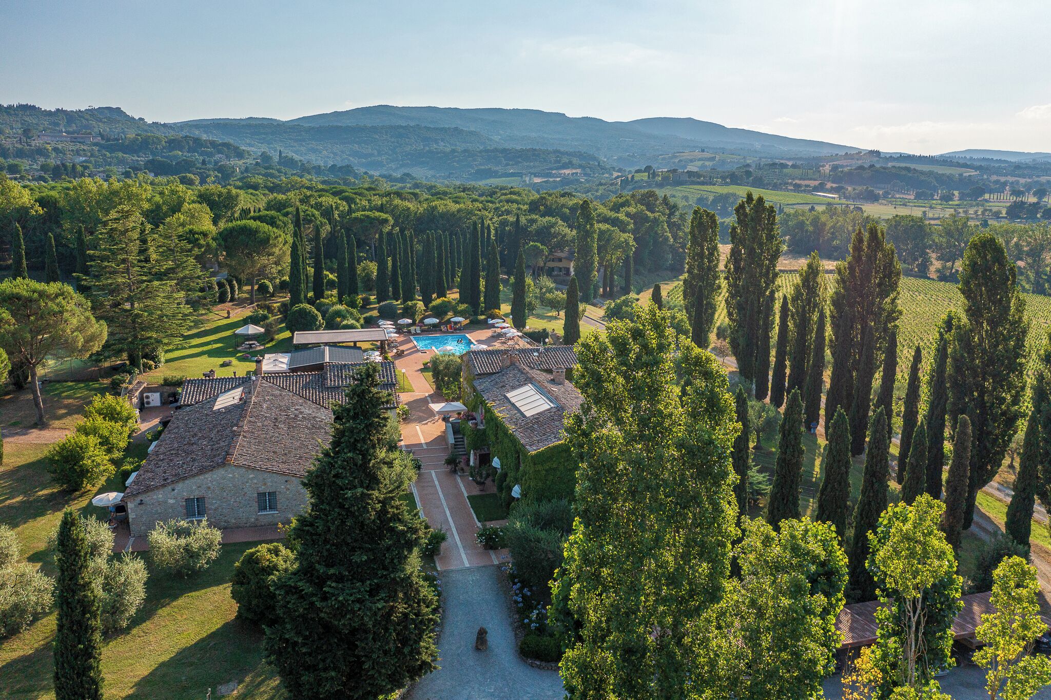 A villa with a swimming pool, surrounded by tall cypress trees and a view of rolling hills.