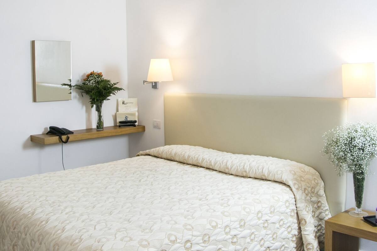 A neatly arranged bedroom with a bed, bedside lamps, a bedside table with a vase, and a mirror on the wall.