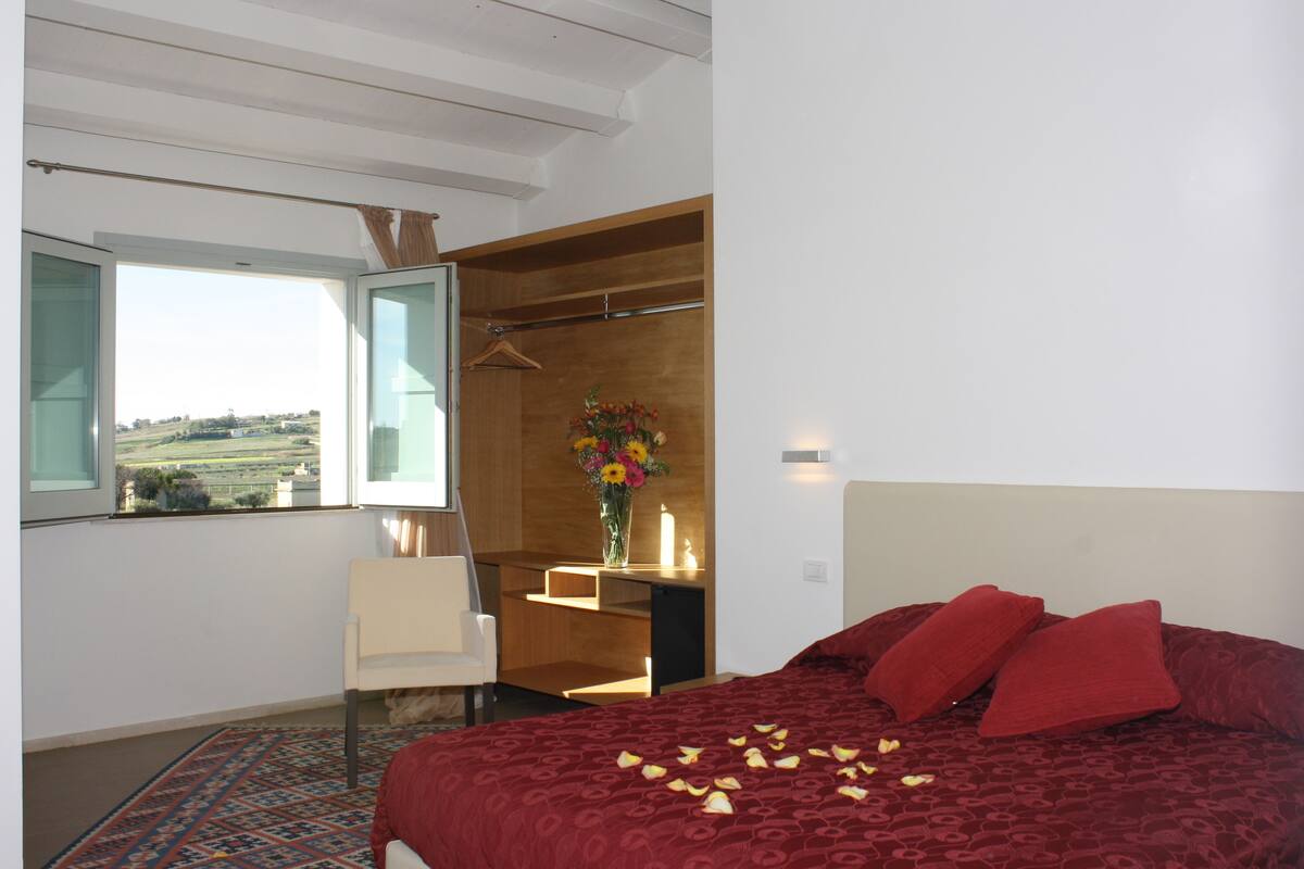 A bedroom with a large bed, a desk with a vase of flowers, and a view of the countryside through a window.