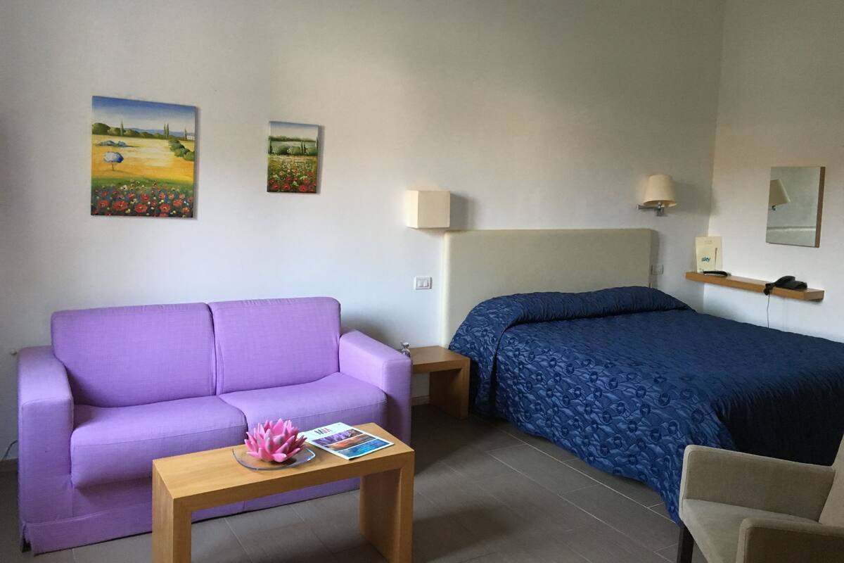A hotel room with a purple sofa, a bed with a blue bedspread, a wooden nightstand, a small table with a magazine and a pink flower, and two wall-mounted lamps.