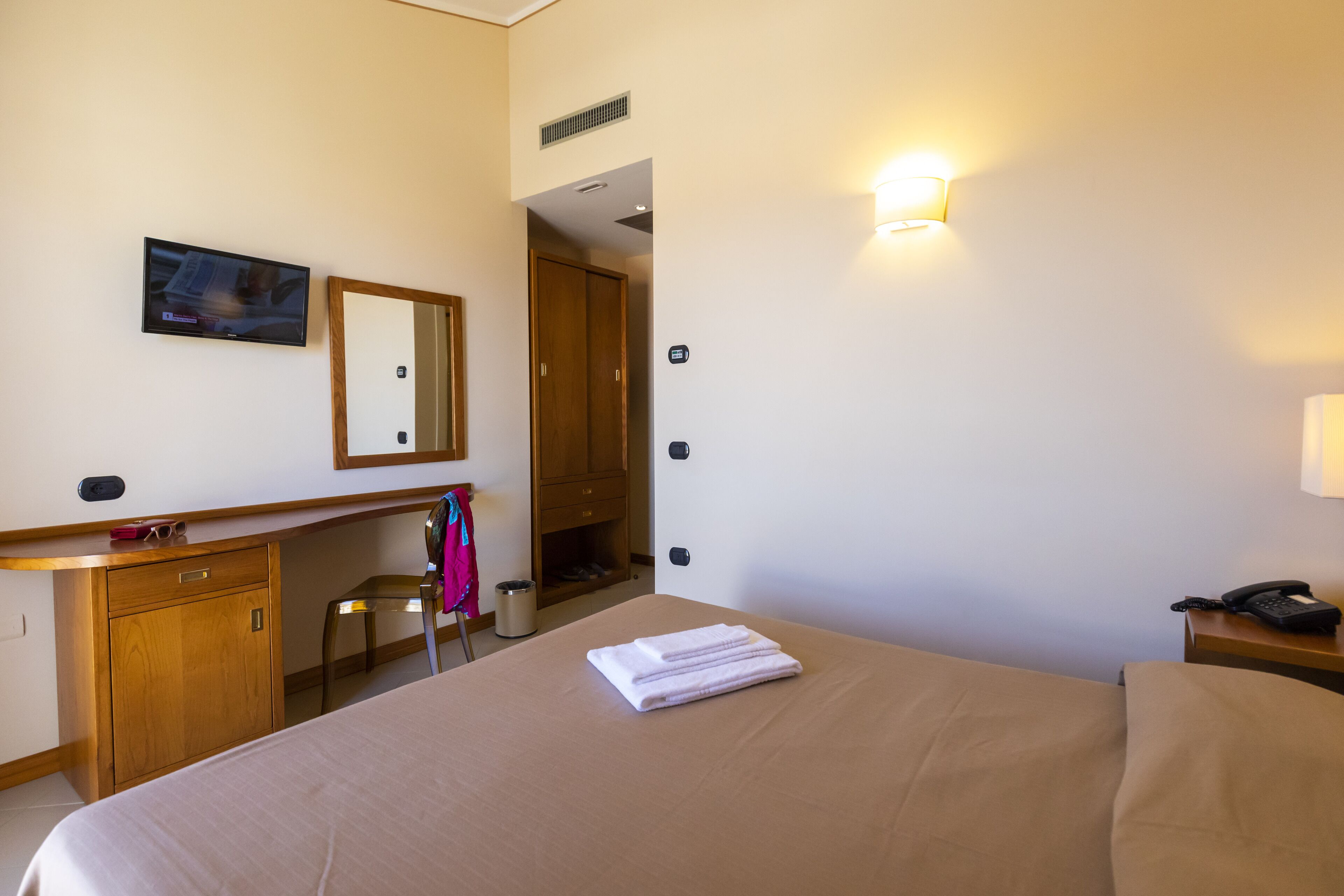 Habitación de hotel con cama, escritorio, silla, espejo, televisor montado en la pared y teléfono en la mesita de noche.