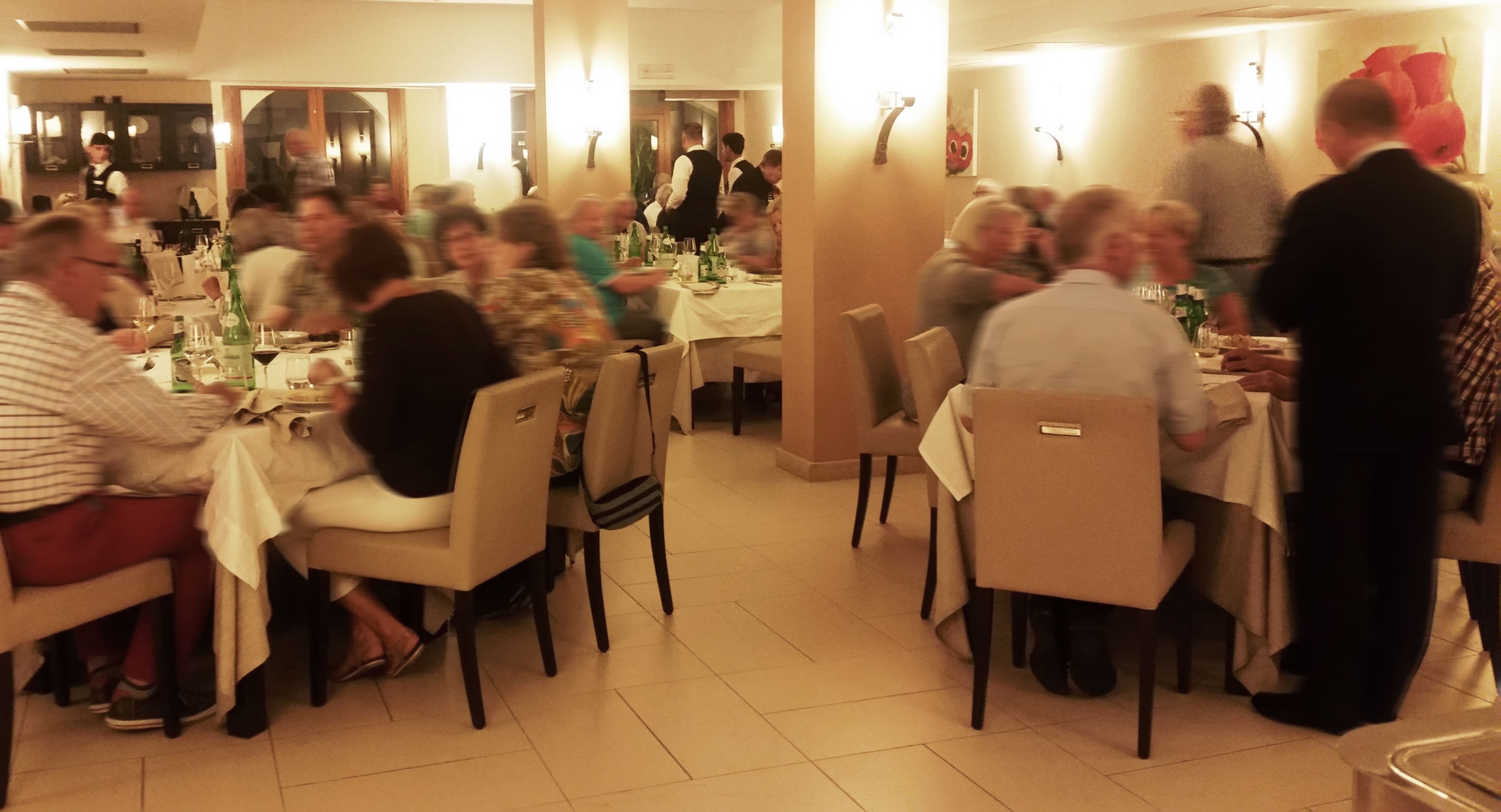 Un comedor con varias mesas y sillas, un camarero con uniforme, y un grupo de personas conversando.