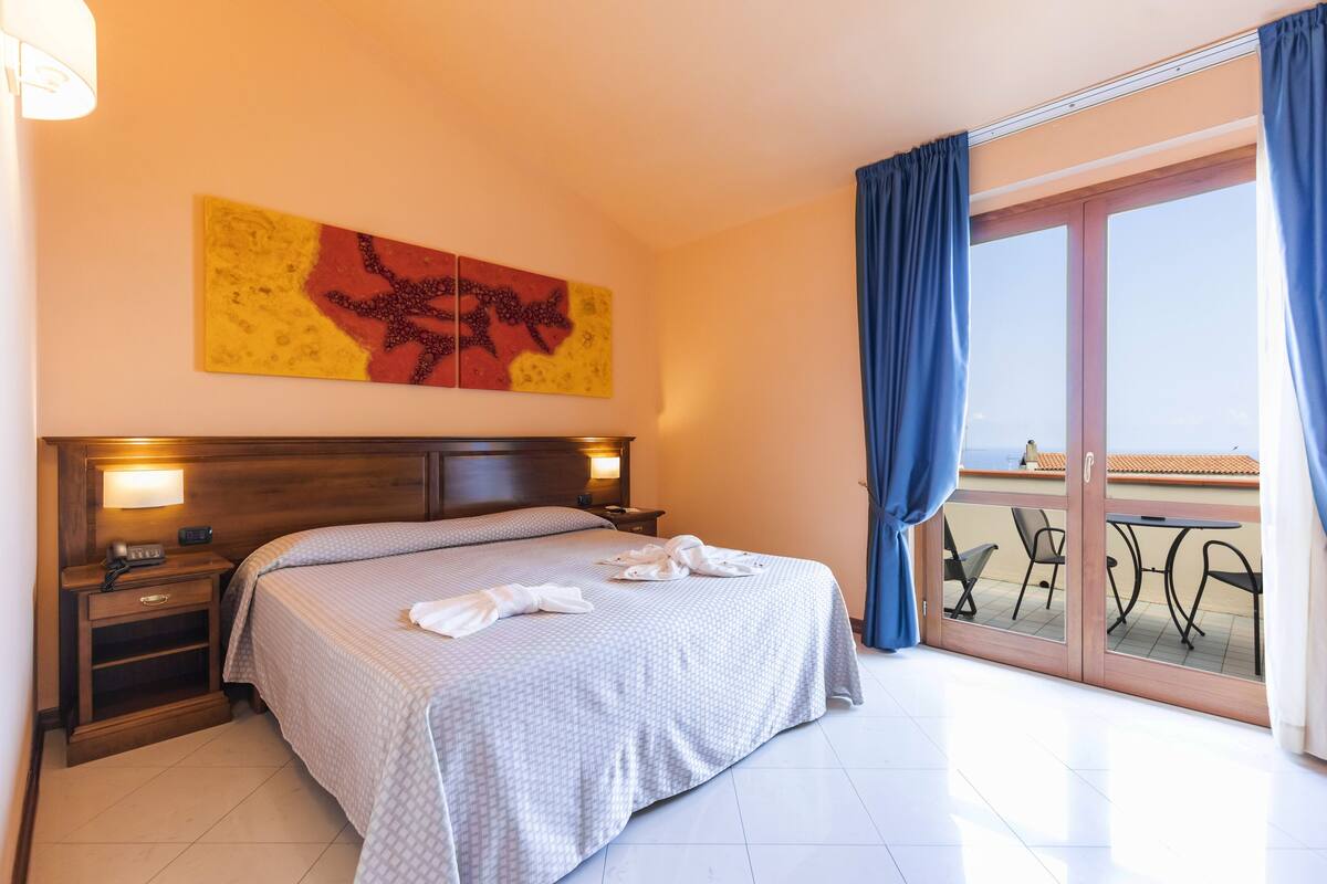 A hotel room with a large bed, a nightstand, a wall-mounted artwork, and a balcony with outdoor furniture.