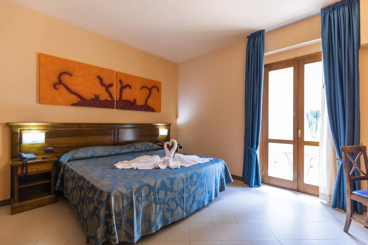 A hotel room with a large bed, a wooden headboard, a nightstand with a lamp, and a wall-mounted artwork.