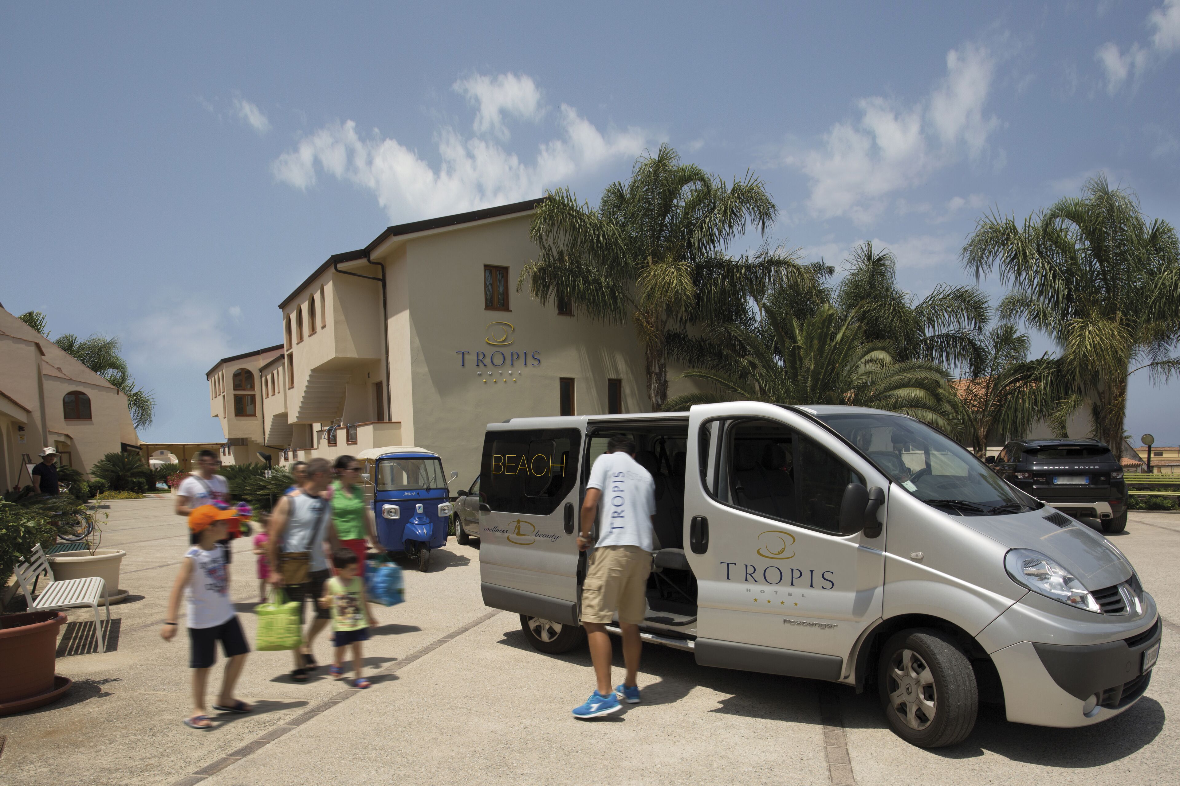 Un fourgon blanc arborant le logo « TROPIS » est garé sur un parking. Des personnes se promènent aux alentours et, au fond, on aperçoit un bâtiment portant l’inscription « TROPIS ».