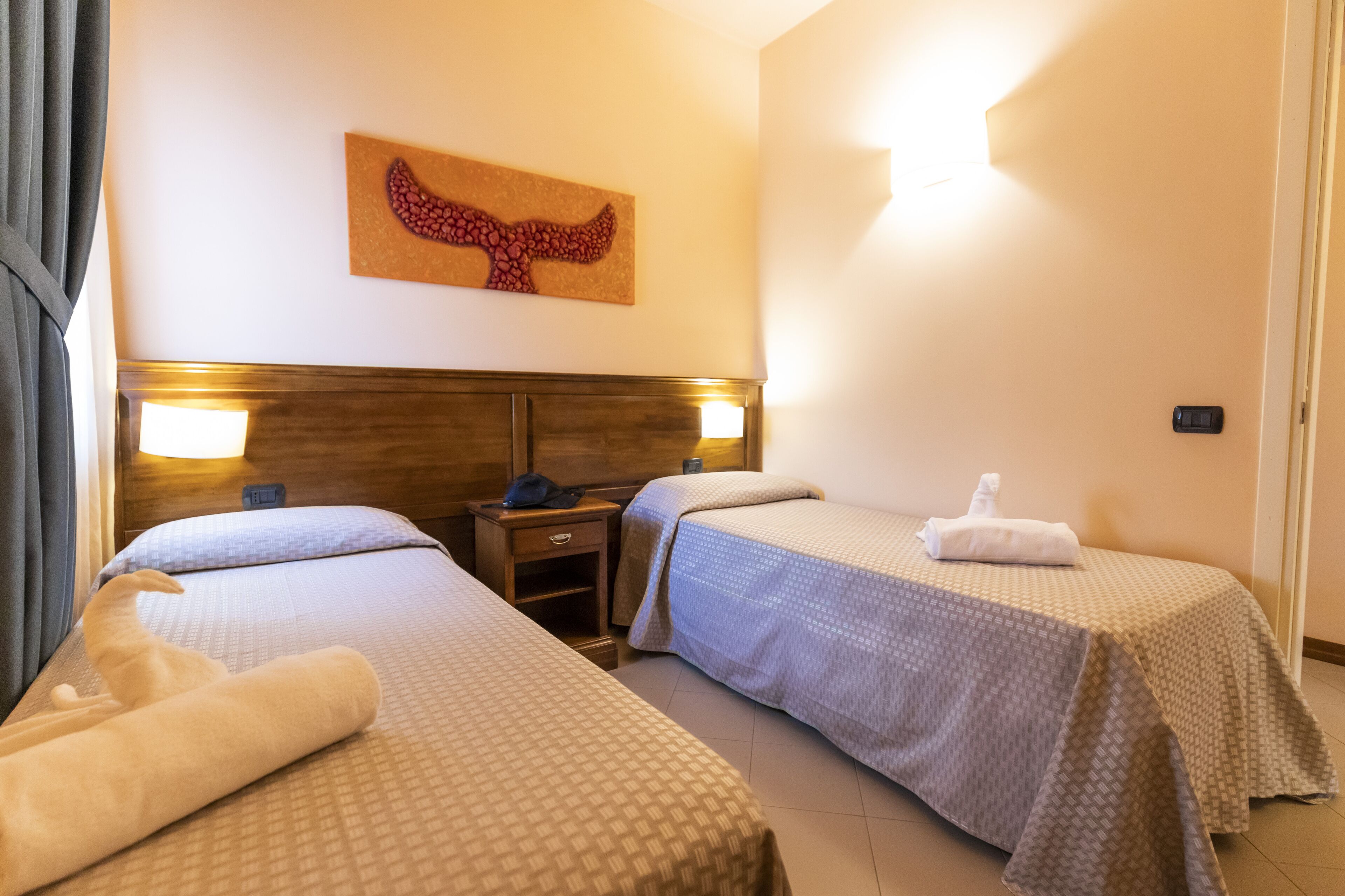 A hotel room with two beds, a wooden headboard, wall-mounted lamps, a clock, and a wall art piece.