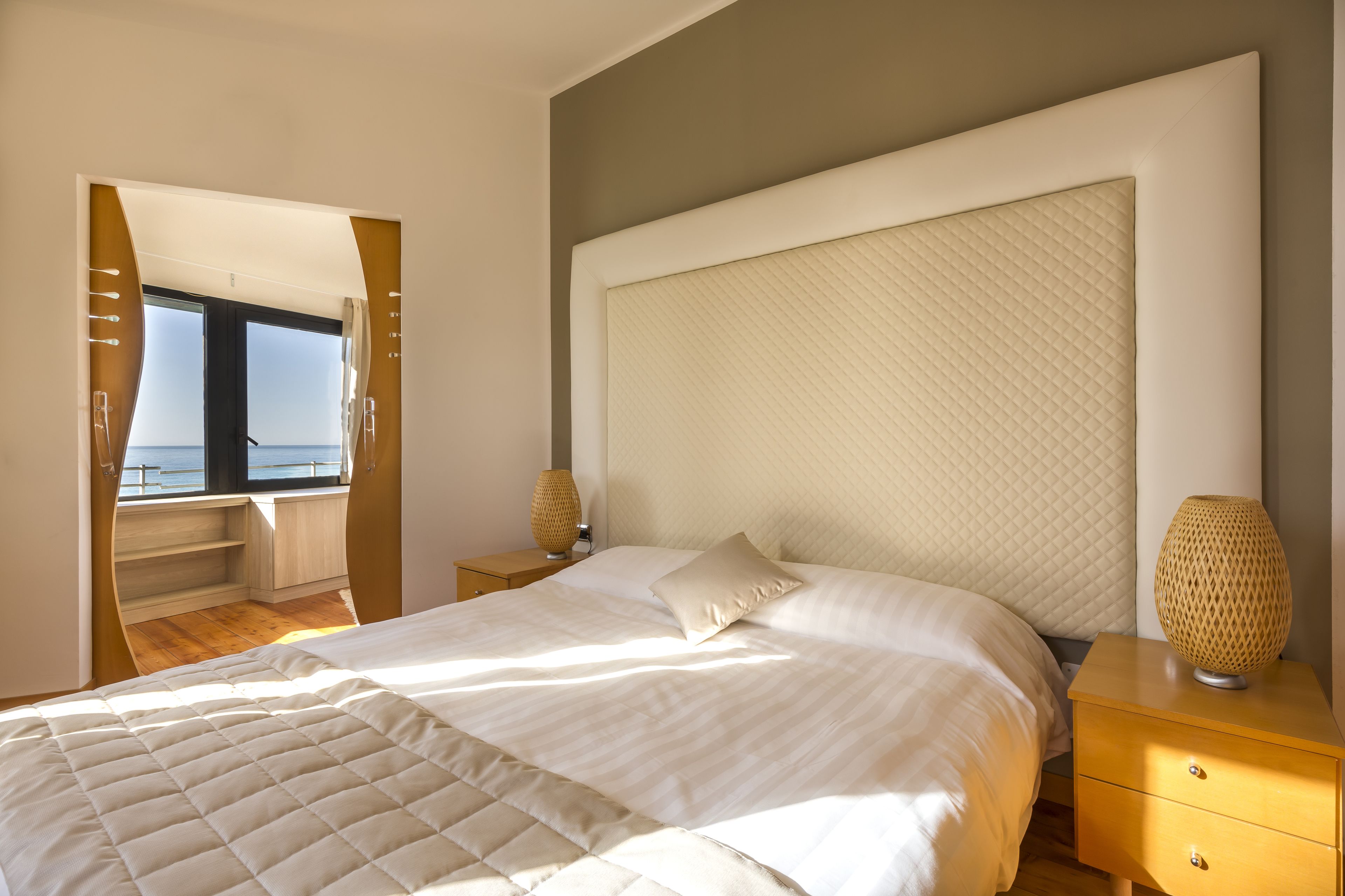 A bedroom with a large bed, a nightstand, a lamp, and a view of the sea through a window.