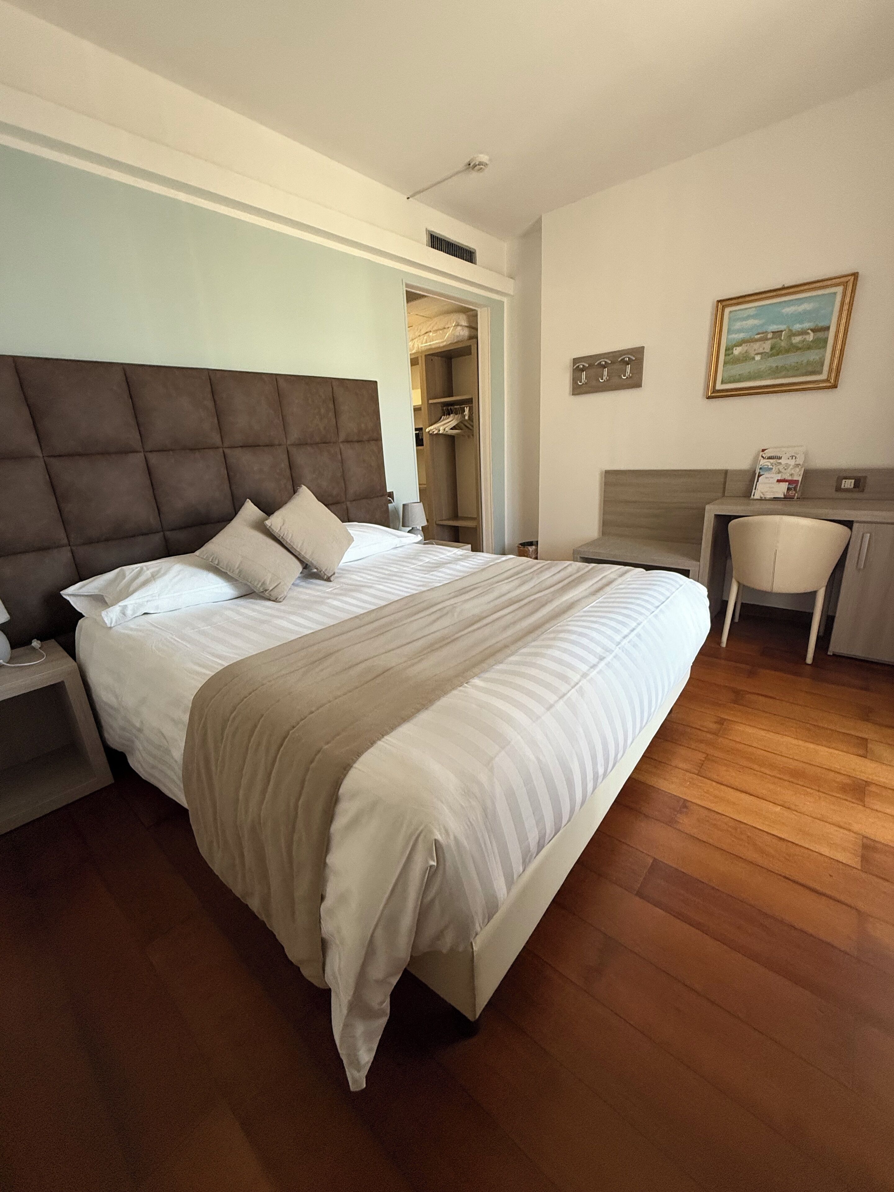 A bedroom with a large bed, a bedside table, a desk with a chair, a wardrobe, and a painting on the wall.