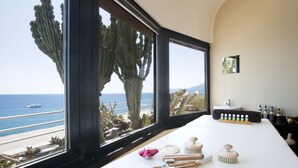 A room with a large window overlooking a beach and ocean, a white table with toiletries, and a view of a cactus outside. - Hotel Parigi & Spa (Bordighera)