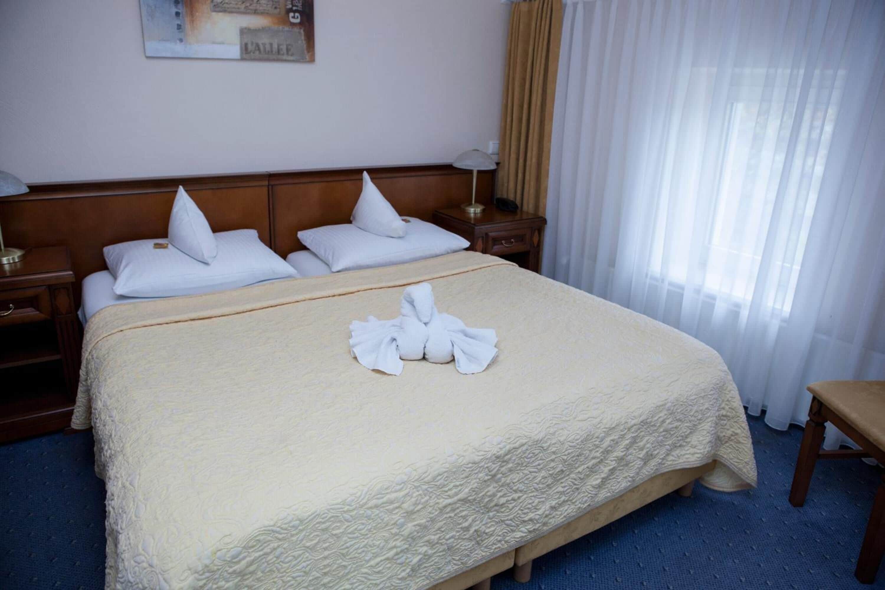 Habitación de hotel con una cama grande, dos mesitas de noche, un banco de madera y un ventanal con cortinas ligeras.