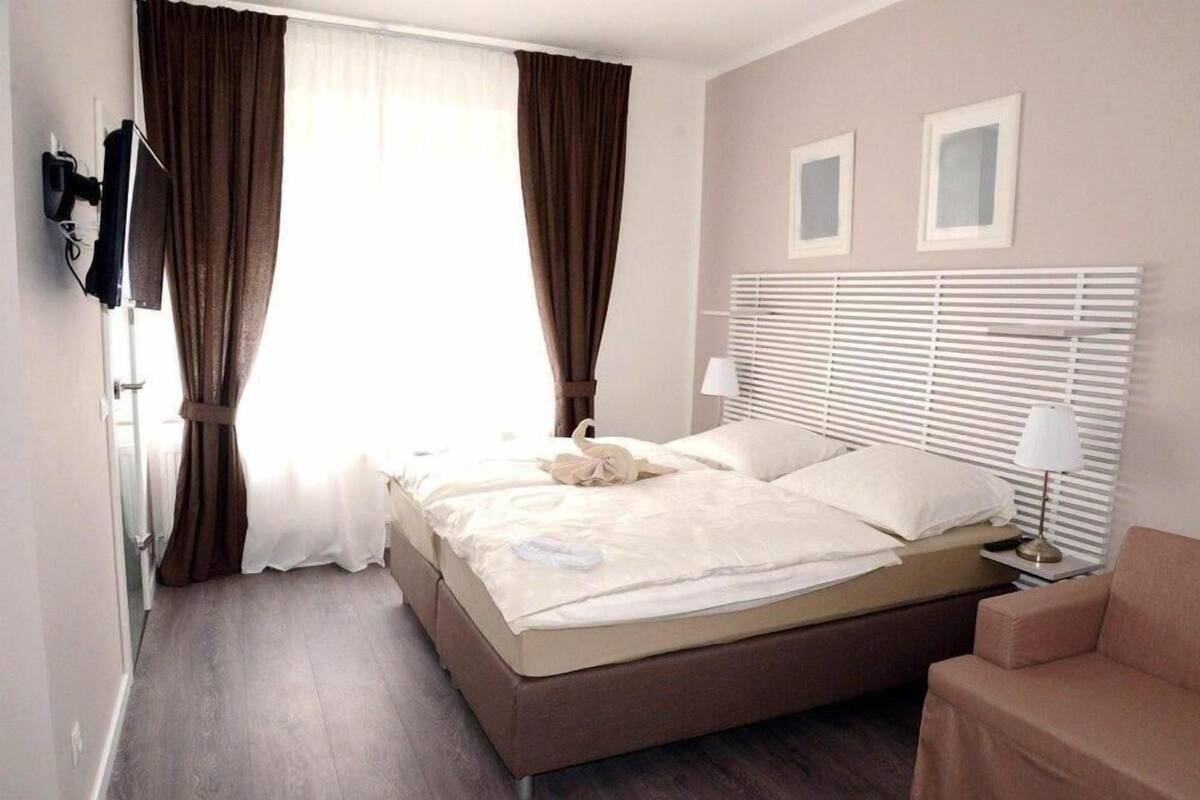 A hotel room with a bed, a bedside table with a lamp, a TV mounted on the wall, and a window with curtains.