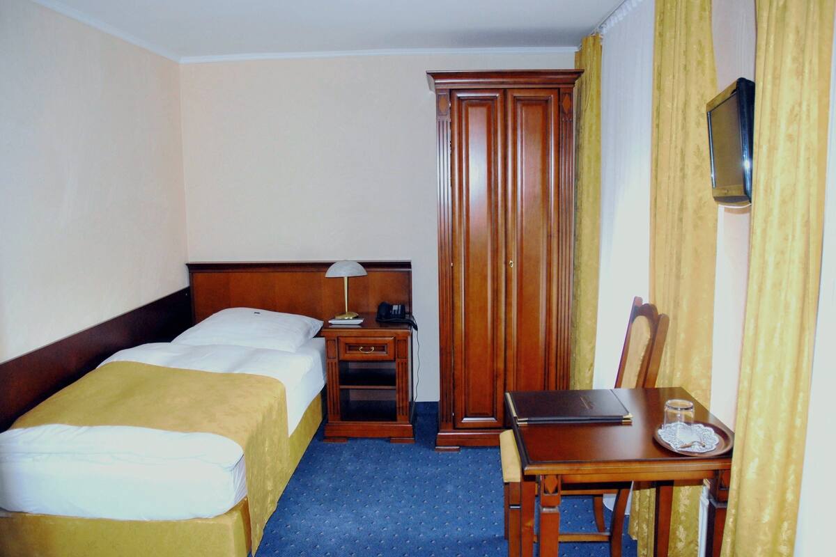 A hotel room with a bed, bedside table, lamp, wardrobe, and a small table with a laptop and a glass.