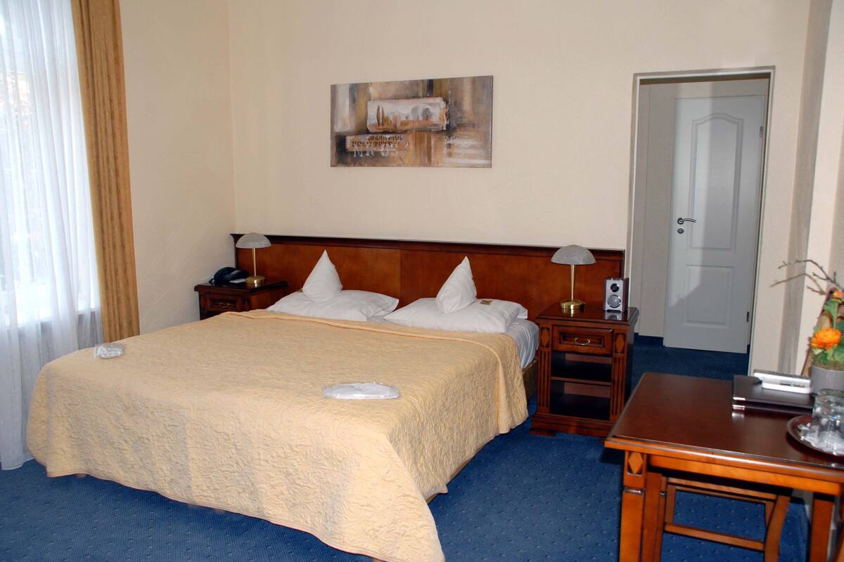 A hotel room with a large bed, two bedside tables with lamps, a wooden nightstand, a desk with a chair, and a door.