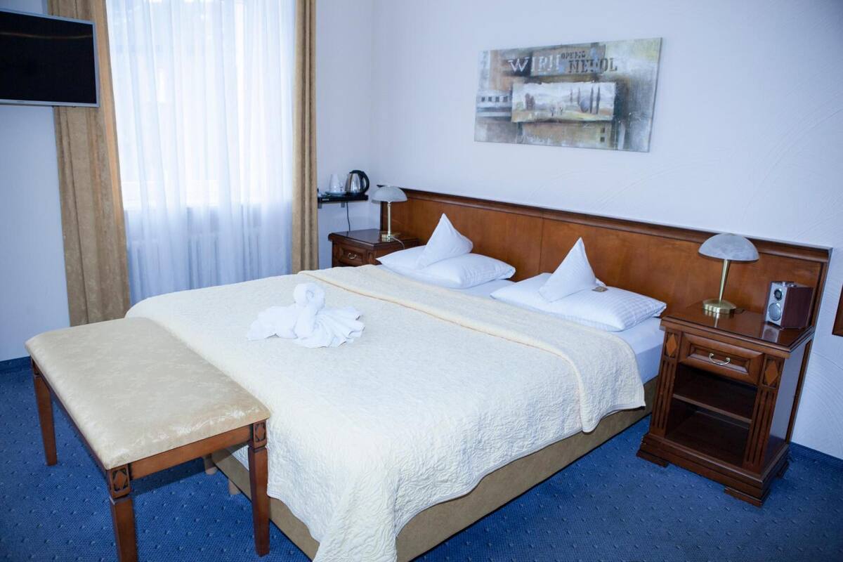 A hotel room with a large bed, a bedside table, a bench, a flat-screen TV mounted on the wall, and a painting above the bed.