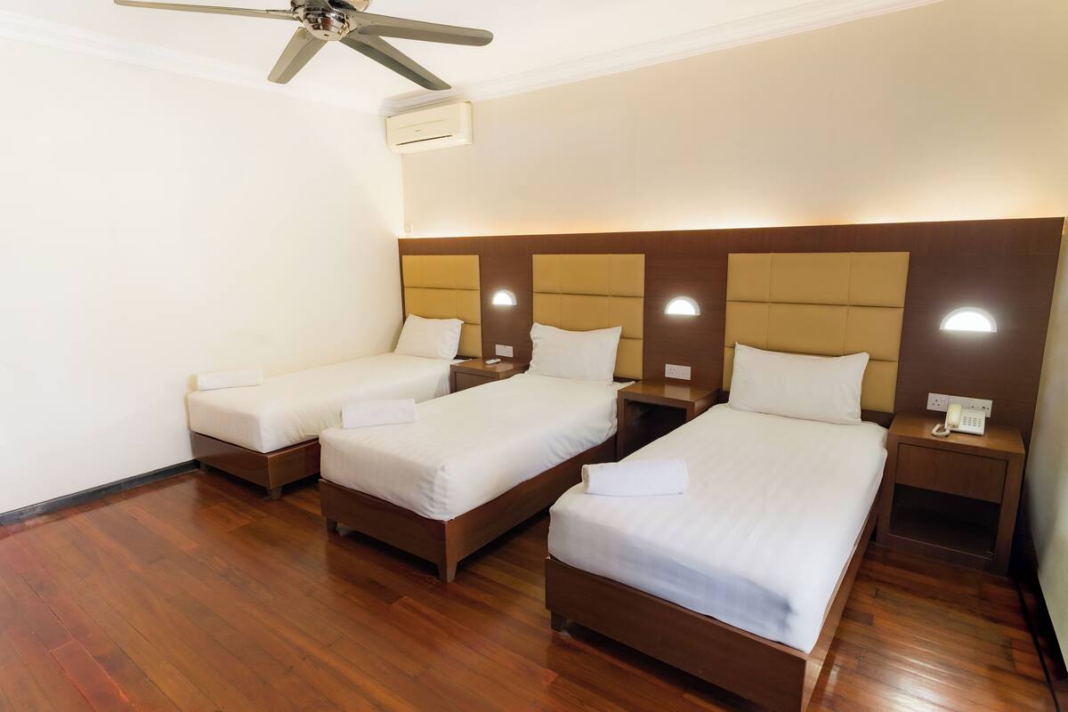 A hotel room with three single beds, wooden flooring, a ceiling fan, and a wall-mounted air conditioner.