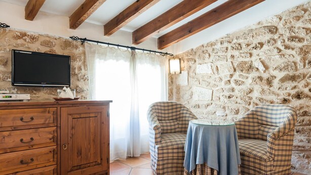 A cozy room with a stone wall, wooden ceiling beams, a TV on a wooden cabinet, and two plaid chairs around a small table. - Can Simo Turismo de Interior (Alcúdia)