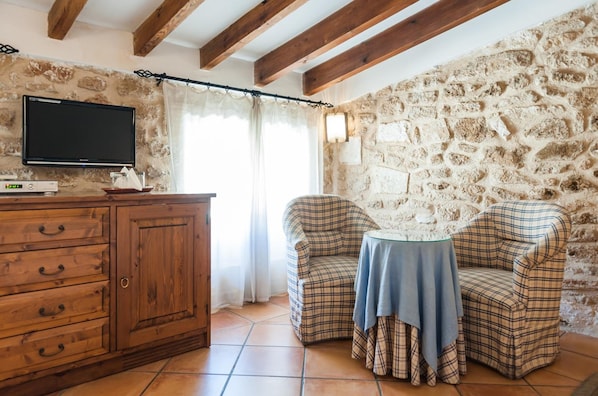 Una habitación acogedora con pared de piedra, vigas de madera en el techo, un televisor sobre un mueble de madera y dos sillones a cuadros alrededor de una mesita.