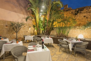 Un área de comedor al aire libre con mesas y sillas, rodeada de plantas y un muro de piedra.
