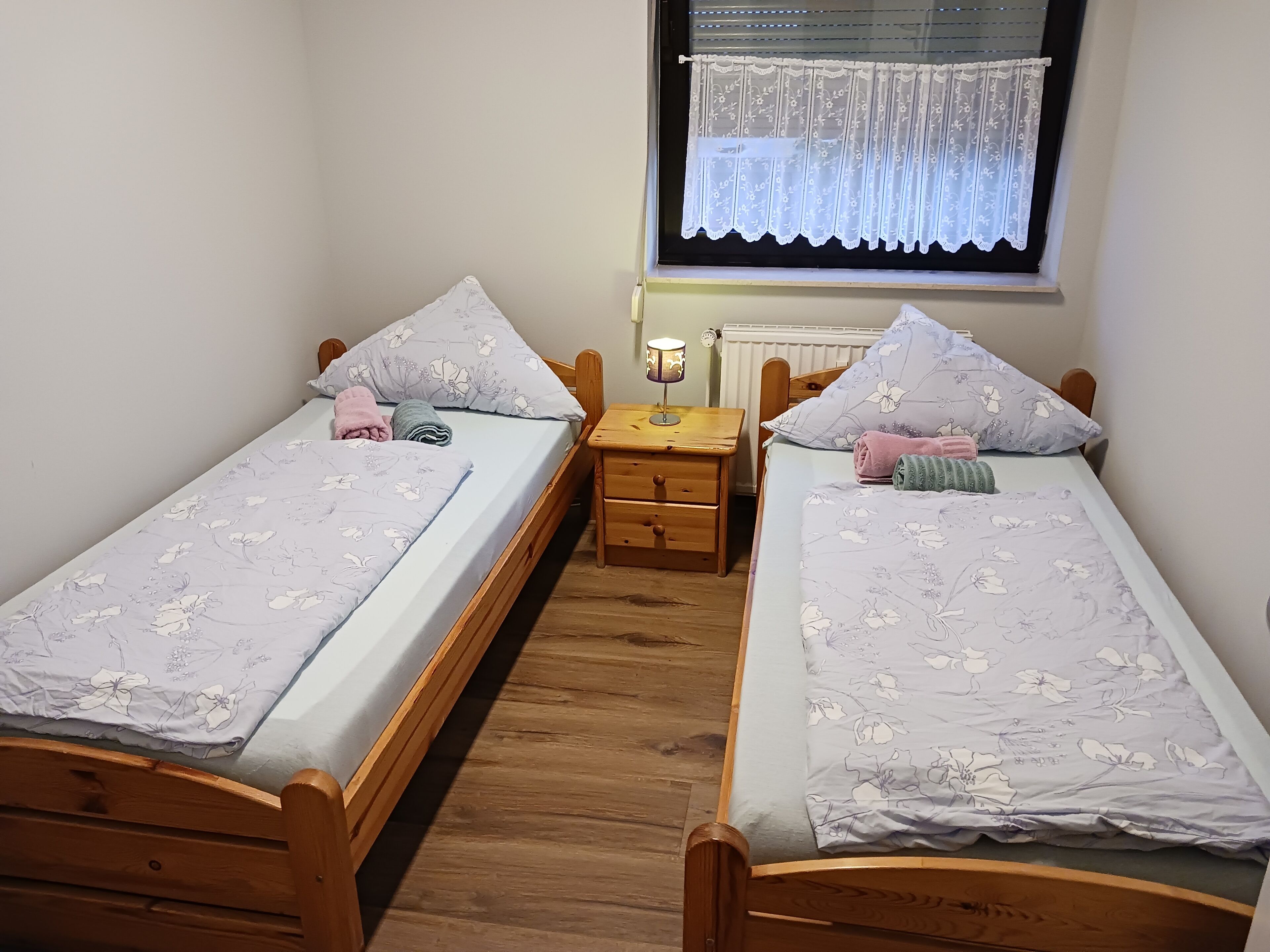 Un dormitorio pequeño y acogedor con dos camas individuales, una mesita de noche con lámpara y una ventana con cortina de encaje.