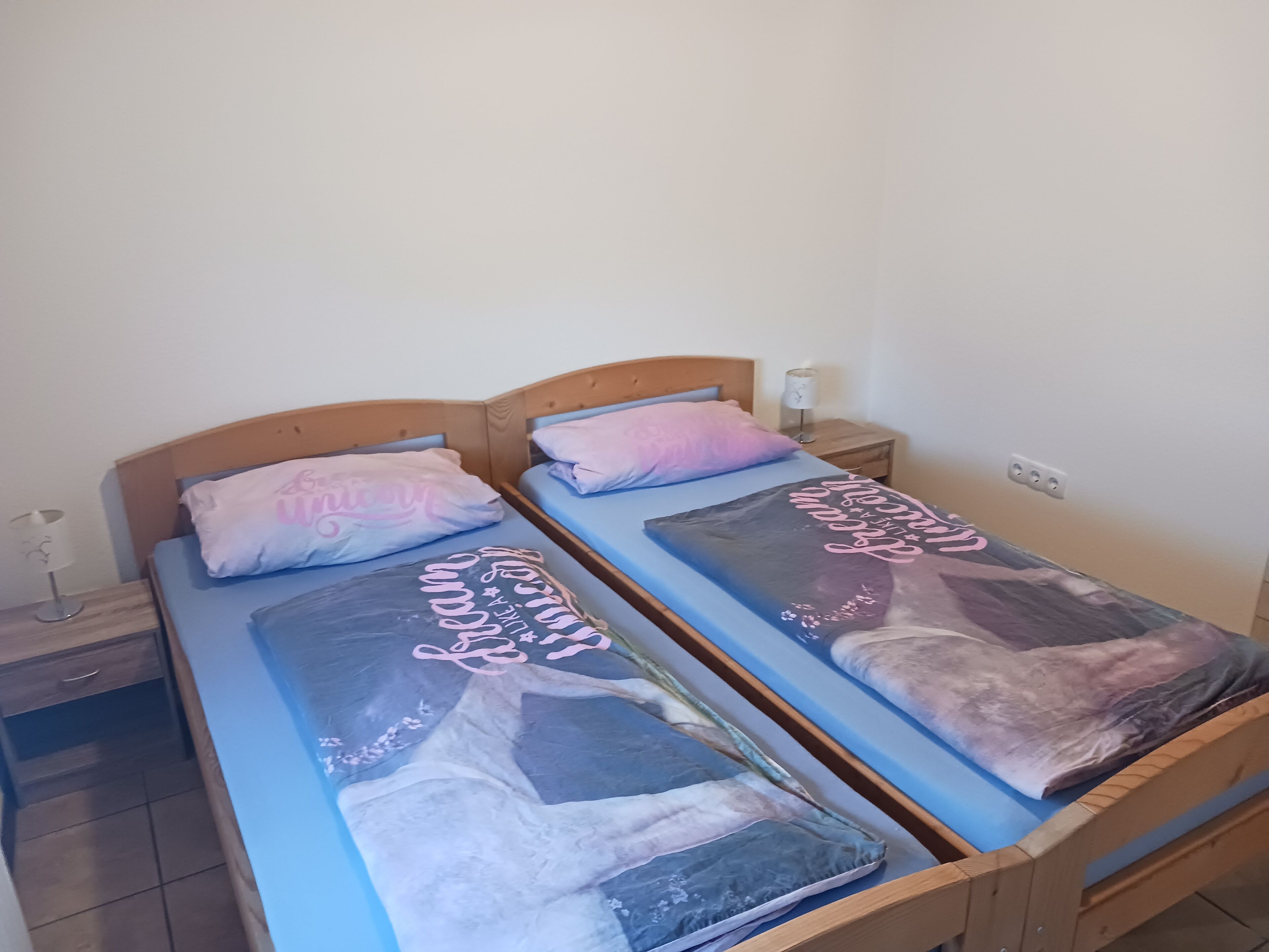 Een tweepersoonsbed met een blauwe matras en een decoratief kussen. Er is een houten hoofdbord, een nachtkastje met een lamp en een stopcontact aan de muur.
