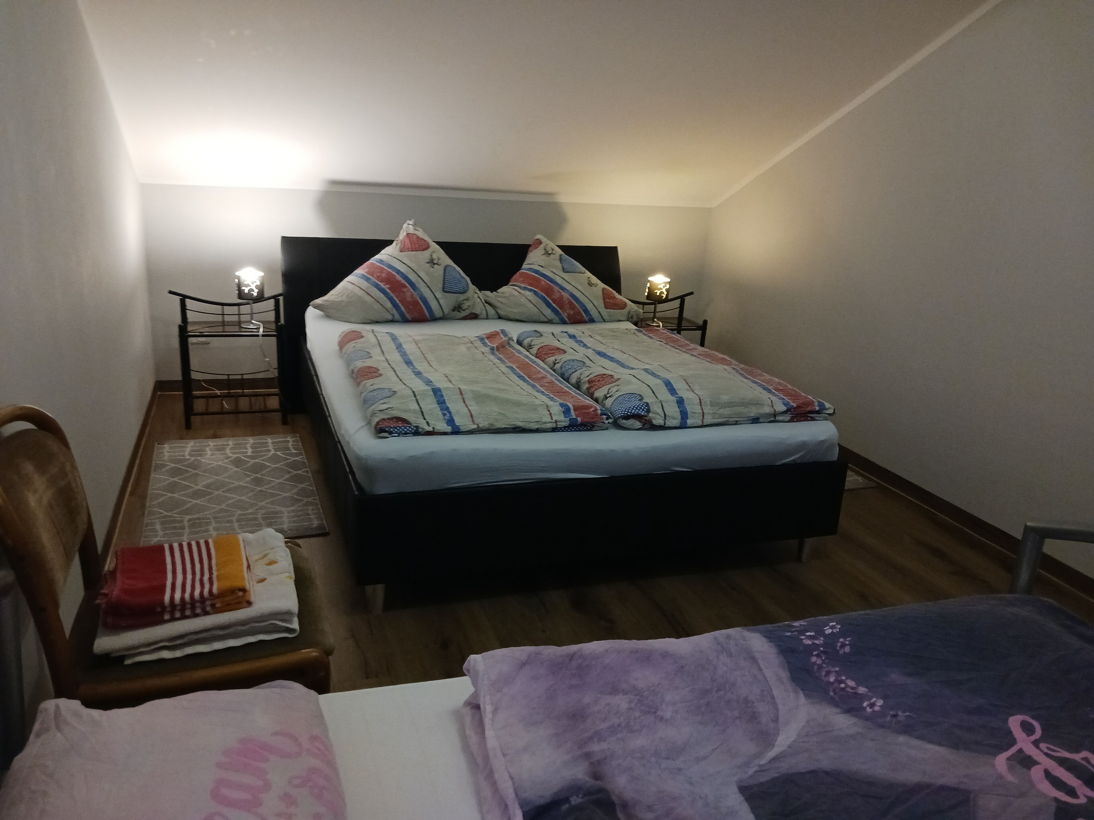 Ein kleines, aufgeräumtes Schlafzimmer mit einem Einzelbett, einem Nachttisch, einem Stuhl und einem Beistelltisch mit Lampe.