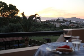 A table with a glass of red wine, a plate with food, and a view of a sunset over a town with buildings and mountains in the background.