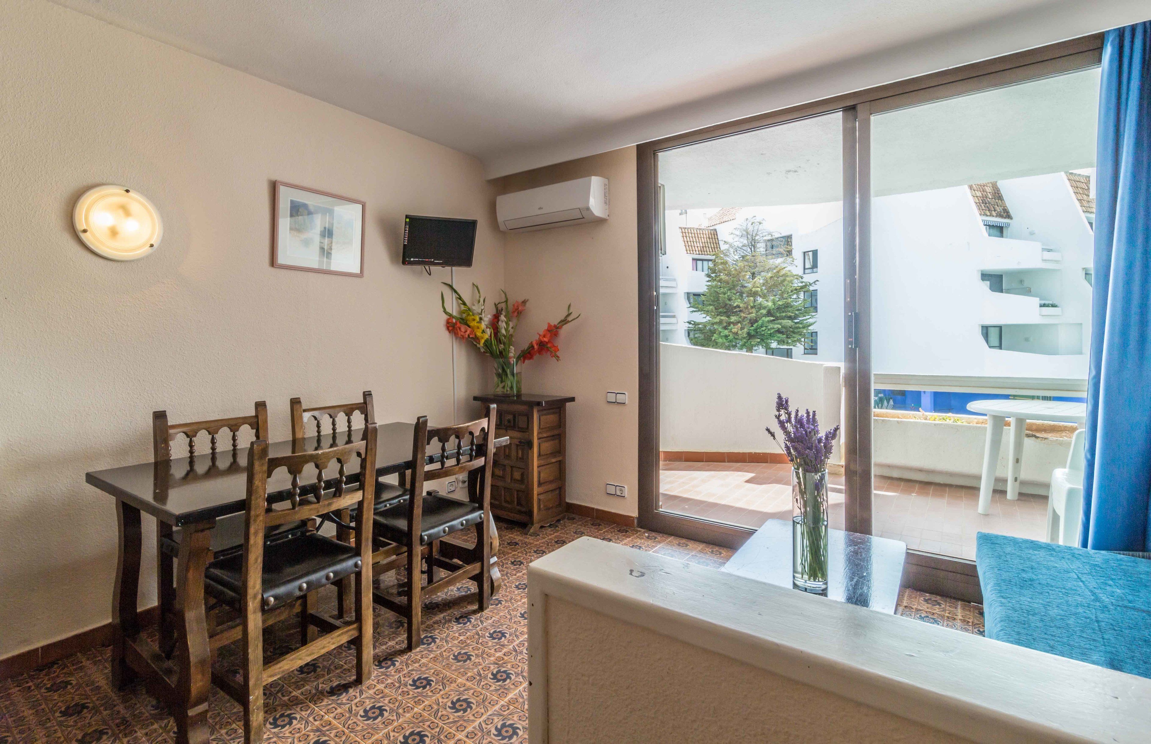 A room with a dining table, chairs, a TV, an air conditioner, a window with a view of buildings, and a vase with flowers.