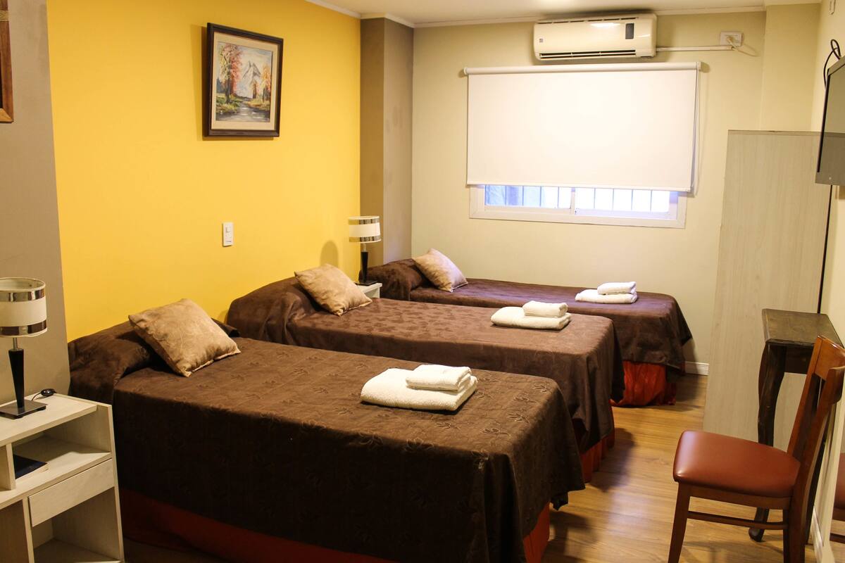 A hotel room with two beds, a desk, a chair, an air conditioner, and a window with blinds.