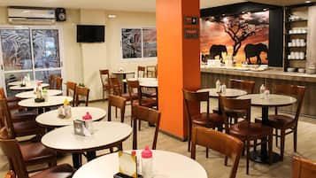 Un restaurante con mesas y sillas redondas, una barra con un mural de cebra y elefante, y un televisor montado en la pared.