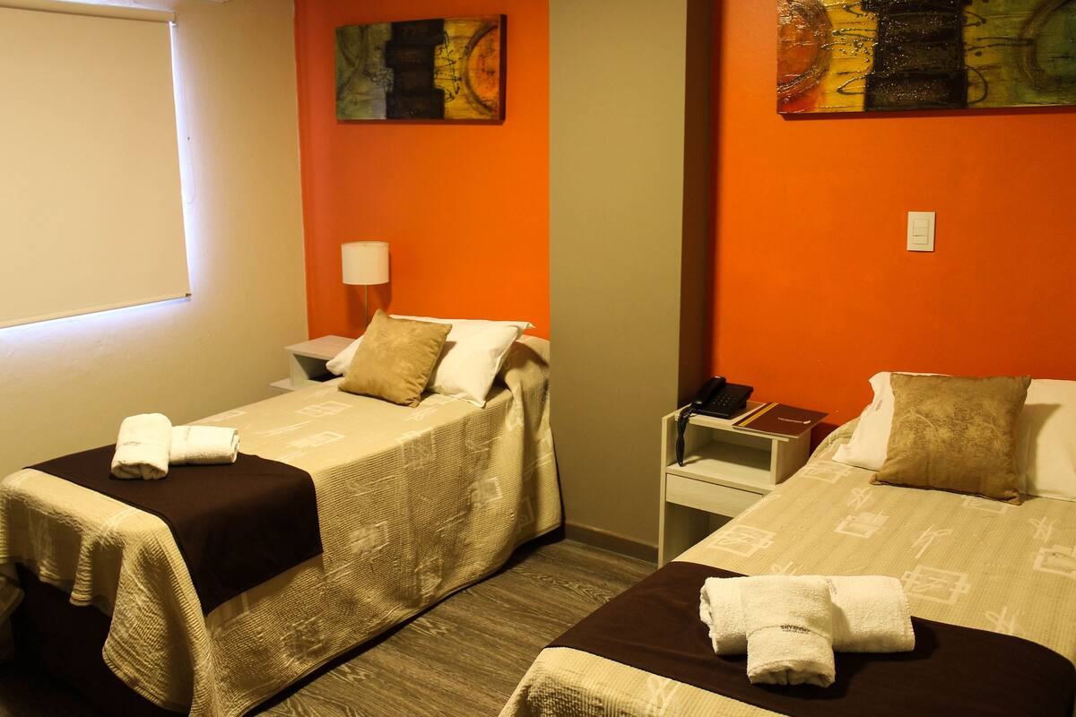 A hotel room with two beds, each with a brown bedspread and white pillows, a nightstand with a lamp, and artwork on the walls.